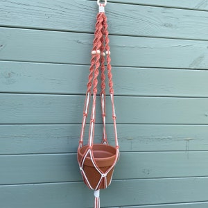 May include: A macrame plant hanger with a terracotta pot, featuring a pink and white rope design with wooden beads.