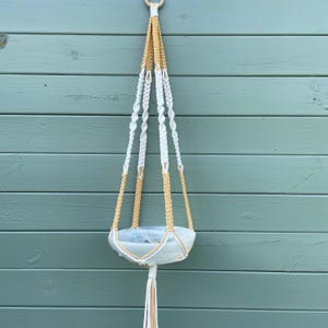 May include: A cream and tan macrame plant hanger, suspended from a wooden ring. The plant holder is a white ceramic bowl. The hanger is made of knotted rope, creating a decorative piece for displaying plants. The plant holder is designed to hold a plant pot.