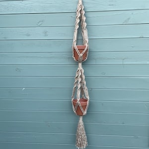 May include: A macrame plant hanger with two terracotta pots hanging from it. The hanger is made of natural coloured string and has a tassel at the bottom.