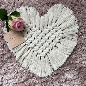 May include: A white heart-shaped macrame coaster with fringe. The coaster is made with cotton cord and has a delicate, handmade look. The coaster is sitting on a plush, pink rug. The text on the tag reads "Made with Love Overley Handmade".