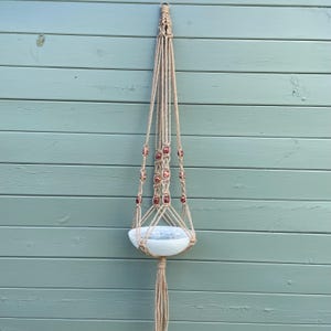May include: A natural-toned macrame plant hanger with red and brown beads, holding a white ceramic bowl. The plant holder is suspended against a light blue wooden wall. The design is suitable for indoor or outdoor use.