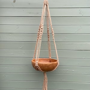 May include: A wooden hanging planter with a natural finish, suspended by beige macrame rope. The planter has a rounded bowl shape and a tassel at the bottom. The background is a light green wooden wall.