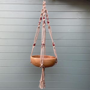 May include: A hanging macrame plant pot holder with a wooden bowl. The beige rope is knotted and decorated with red beads. The bowl is made of light-coloured wood. The hanger is against a light blue-green background.