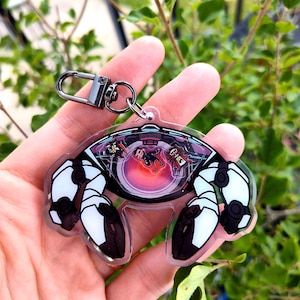 Puede incluir: Un llavero acrílico transparente con un diseño en blanco y negro. El llavero presenta un diseño circular con un centro rojo y naranja, y una escena con figuras en el interior. Tiene un cierre plateado.