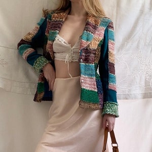 May include: A multi-coloured patchwork jacket with a teal base colour. The jacket has a button closure and a collar. The model is wearing a white lace bralette and a light pink skirt.