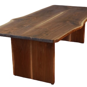 May include: A large, live edge walnut wood dining table with a natural, brown finish. The table has a rectangular top with a live edge on each side. The table is supported by two rectangular legs.