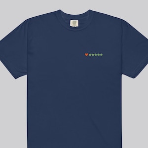 Pode incluir: Uma t-shirt azul-marinho de manga curta com um pequeno gráfico bordado no peito esquerdo. O design consiste num ícone de coração laranja seguido por cinco pequenas estrelas verdes dispostas numa linha horizontal no tecido azul escuro.