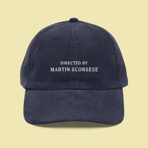 Könnte beinhalten: Eine marineblaue Baseballkappe aus Cord mit weiß gesticktem Text, der "Regie: Martin Scorsese" lautet.