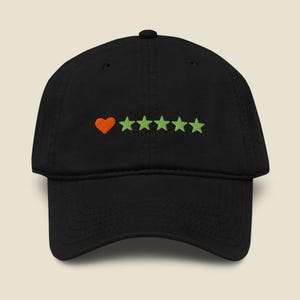 Pode incluir: Um boné de beisebol preto com um design bordado no painel frontal. O bordado consiste num coração laranja seguido por cinco estrelas verde-claras dispostas numa linha horizontal no centro do boné.