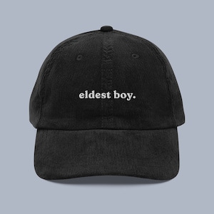 Op de afbeelding: Een zwarte corduroy baseballpet met witte geborduurde tekst die "eldest boy." luidt.