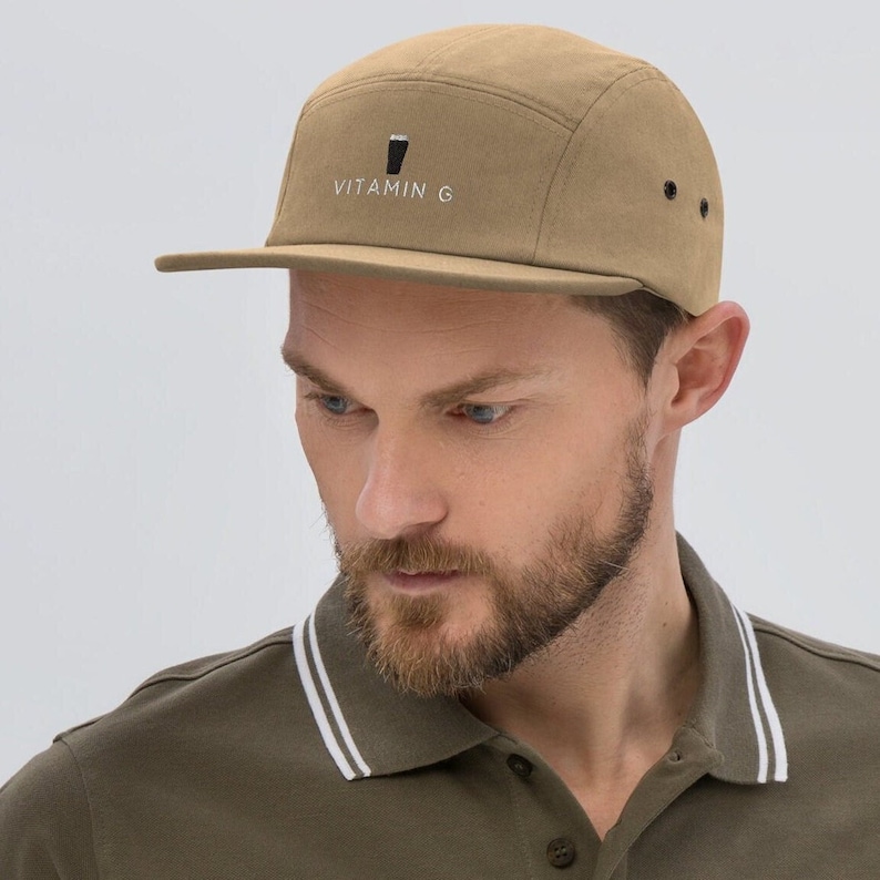 K&ouml;nnte beinhalten: Eine beige 5-Panel-Kappe mit einem schwarz-wei&szlig;en Logo, auf dem vorne "VITAMIN G" steht.