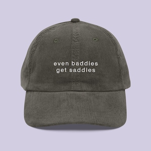 May include: An olive green corduroy baseball cap with the white text "even baddies get saddies". The cap has a curved peak and an adjustable strap. A casual hat for everyday wear.