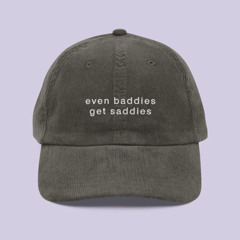 May include: Olive green corduroy baseball cap with the text "even baddies get saddies" in white. The cap has a curved brim and an adjustable strap at the back. The hat is a casual accessory.