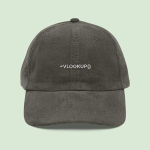 May include: A dark olive green corduroy baseball cap with a curved brim. The front of the cap features the white text "=VLOOKUP()". The cap has a structured crown and an adjustable strap at the back.
