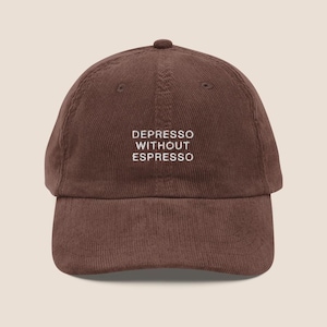 May include: A brown corduroy baseball cap featuring the embroidered white text DEPRESSO WITHOUT ESPRESSO centred on the front panel. The hat has a structured crown, a curved brim, and a button at the top, showcasing a textured fabric finish.