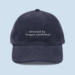 May include: A navy blue corduroy baseball cap with white embroidered text that reads "directed by Yorgos Lanthimos".