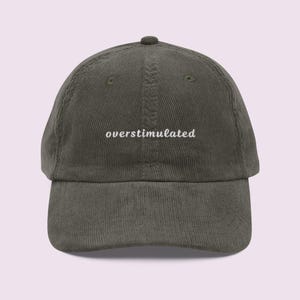 May include: A corduroy baseball cap in a muted olive green color. The cap features the word "overstimulated" embroidered in white, centered on the front. The hat has a curved brim and an adjustable strap.