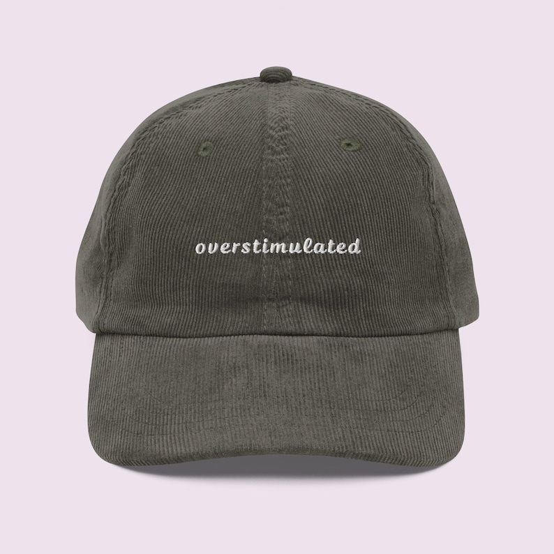 May include: A corduroy baseball cap in a muted olive green color. The cap features the word "overstimulated" embroidered in white, centered on the front. The hat has a curved brim and an adjustable strap.