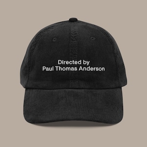 May include: Black corduroy baseball cap featuring the text "Directed by Paul Thomas Anderson" in white. The cap has a curved peak and a rounded crown. A stylish accessory for film fans.