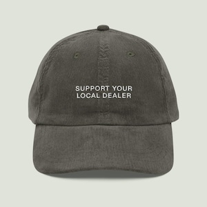 Pode incluir: Um boné de beisebol de veludo cotelê cinza-escuro com o texto branco bordado SUPPORT YOUR LOCAL DEALER centralizado no painel frontal. O boné possui uma copa estruturada, aba curva e um botão no topo, exibindo um tecido texturizado.