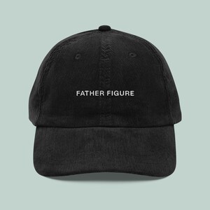 May include: Black corduroy baseball cap with the text "FATHER FIGURE" embroidered in white. The cap has a curved brim and a rounded crown. A casual accessory for everyday wear.