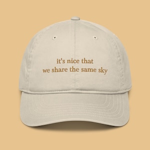 May include: A beige baseball cap with the text "it's nice that we share the same sky" embroidered in brown on the front.