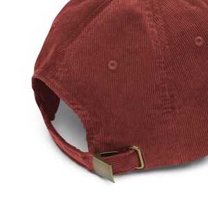 May include: A deep red corduroy baseball cap featuring a textured fabric surface with vertical ridges. The back of the cap includes an adjustable strap with a metallic brass-toned buckle closure and matching eyelets for ventilation on the crown.