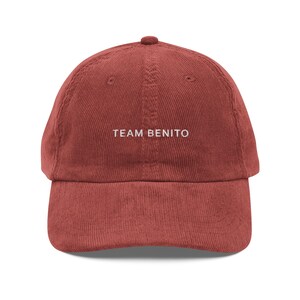 Puede incluir: Gorra de b&eacute;isbol de pana rojo ladrillo con visera curva. El texto "TEAM BENITO" est&aacute; bordado en blanco en la parte delantera. La gorra tiene una corona redondeada y un peque&ntilde;o bot&oacute;n en la parte superior.