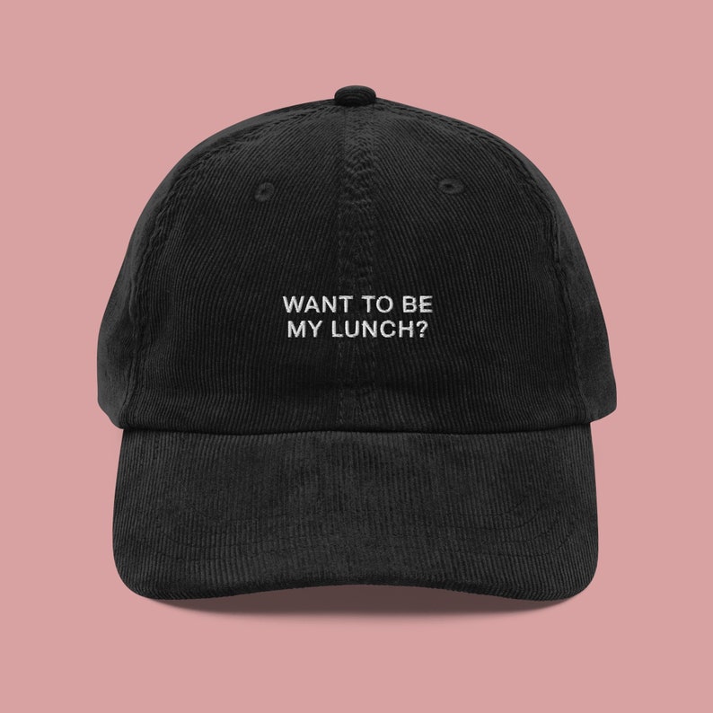 May include: Black corduroy baseball cap with the embroidered text "WANT TO BE MY LUNCH?" in white. The cap has a curved brim and a top button. The background is a solid light pink.