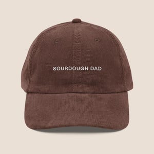 May include: A brown corduroy baseball cap featuring the text SOURDOUGH DAD embroidered in white thread across the front centre. The cap has a structured crown, a curved brim, and a button at the top, showcasing a classic six-panel design.