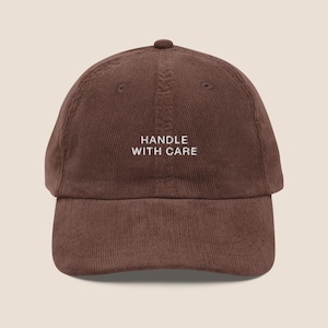May include: A dark brown corduroy baseball cap featuring the embroidered white text "HANDLE WITH CARE" centred on the front panel. The hat has a structured crown, a curved brim, and a button at the top, showcasing a textured fabric finish.