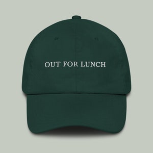 Peut inclure: Une casquette de baseball vert foncé avec le texte "OUT FOR LUNCH" brodé en blanc sur le devant.