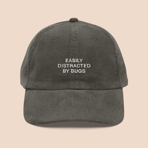 Puede incluir: Una gorra de béisbol de pana verde oscuro con bordado blanco que dice "EASILY DISTRACTED BY BUGS".