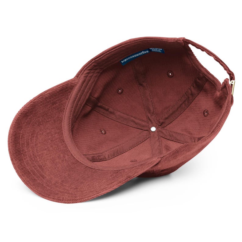 May include: A maroon corduroy baseball cap shown from an interior perspective, featuring a six-panel construction with internal seams, a fabric sweatband, and a blue brand label reading bigaccessories with 100% cotton text, plus a metal buckle.
