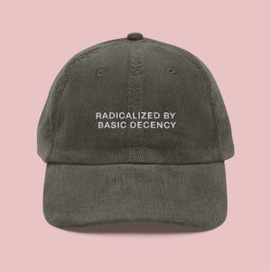 Op de afbeelding: Een donker olijfgroene corduroy baseballcap met de tekst "RADICALIZED BY BASIC DECENCY" in witte blokletters. De cap heeft een gebogen klep en een kleine knoop bovenop. De achtergrond is lichtroze.