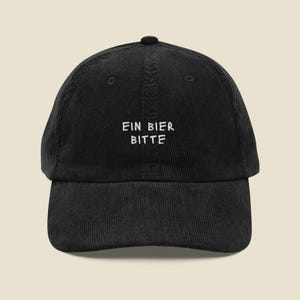 以下が含まれることがあります： フロントパネルの中央に白い刺繍で「EIN BIER BITTE」と施された、黒いコーデュロイ素材のベースボールキャップ。構造化されたクラウン、カーブしたつば、通気用のステッチ入りアイレットを備えた、クラシックな外観の帽子です。