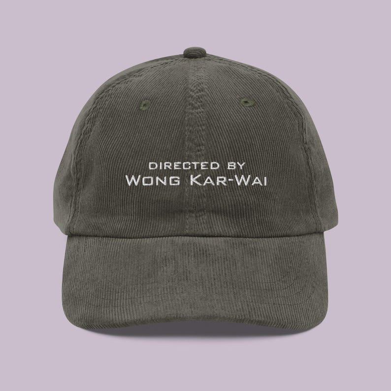 以下が含まれることがあります： 前面に白い刺繍で「DIRECTED BY WONG KAR-WAI」と刺繍された、濃い緑色のコーデュロイ製のベースボールキャップです。