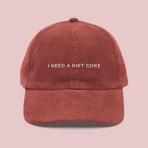 May include: A rust-colored corduroy baseball cap with the text "I NEED A DIET COKE" embroidered in white. The cap has a curved brim and a rounded top, suitable for casual wear.