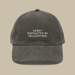 Könnte beinhalten: Olivgrüne Cord-Baseballkappe mit dem gestickten Text "EASILY DISTRACTED BY HELICOPTERS" in Weiß. Die Kappe hat einen gebogenen Schirm und eine runde Krone, ideal für den Alltag.