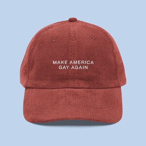 Könnte beinhalten: Eine rote Baseballkappe aus Cord mit dem aufgestickten Text "Make America Gay Again" in weiß.