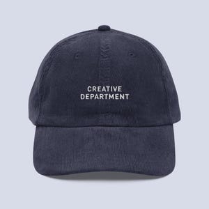 Puede incluir: Gorra de béisbol de pana azul marino con el texto "CREATIVE DEPARTMENT" bordado en blanco. La gorra tiene una visera curva y una corona redondeada. Un accesorio de moda.