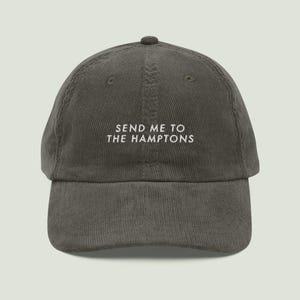 May include: A dark grey corduroy baseball cap featuring the embroidered white text SEND ME TO THE HAMPTONS centered on the front panel. The hat has a structured crown, a curved brim, and a button at the top, showcasing a textured fabric finish.