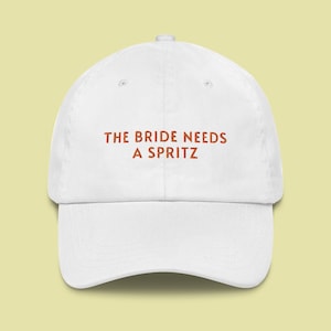 Op de afbeelding: Witte baseballpet met de tekst "THE BRIDE NEEDS A SPRITZ" geborduurd in oranje.