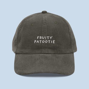 May include: A corduroy baseball cap in a muted olive green color. The cap features the embroidered text "FRUITY PATOOTIE" in white, centered on the front. The hat has a curved brim and an adjustable strap.