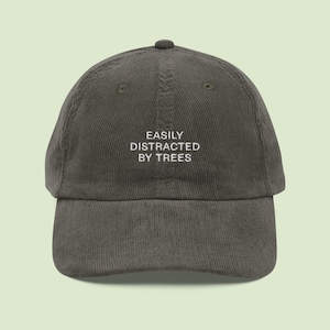 May include: A dark olive green corduroy baseball cap with a curved brim. The cap features the white embroidered text "EASILY DISTRACTED BY TREES" on the front. The hat has a rounded crown and an adjustable strap at the back.