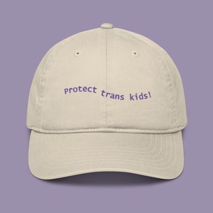 Può includere: Un berretto da baseball beige con il testo "protect trans kids!" ricamato in viola.