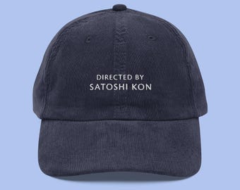 Może przedstawiać: Czapka bejsbolowa z niebieskiego, prążkowanego materiału typu corduroy z białym haftem, na którym widnieje napis "DIRECTED BY SATOSHI KON".