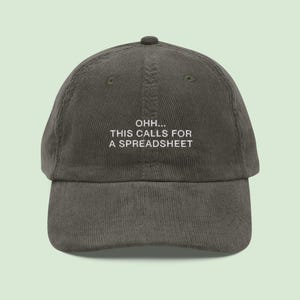 May include: A corduroy baseball cap in a muted olive green color. The cap features the phrase "OHH... THIS CALLS FOR A SPREADSHEET" embroidered in white block letters across the front. The cap has a curved brim and a top button.
