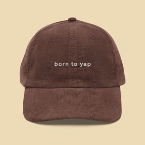 May include: A brown corduroy baseball cap with white embroidery that reads "born to yap".