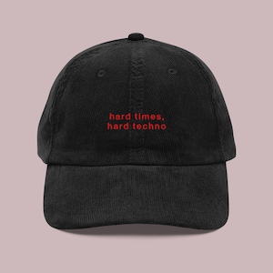 May include: Black corduroy baseball cap with the red text "hard times, hard techno" embroidered on the front. The cap has a curved peak and a button on top.